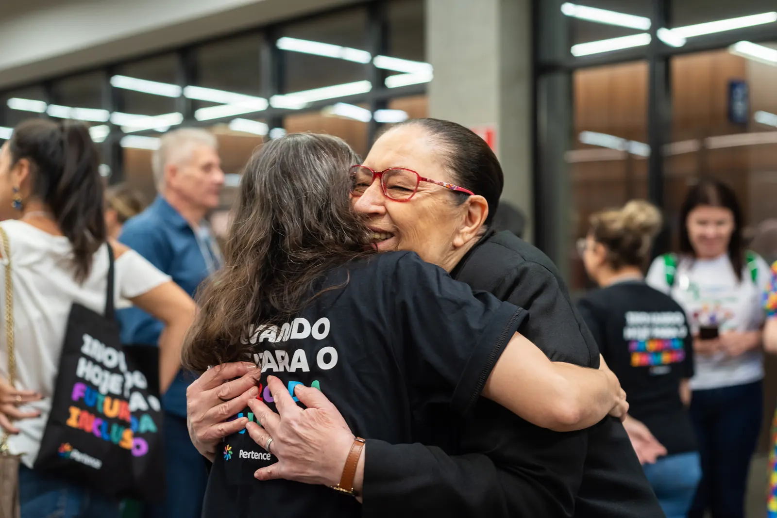 Duas mulheres se abraçam sorrindo em um evento do Pertence; uma delas veste a camiseta do Instituto.